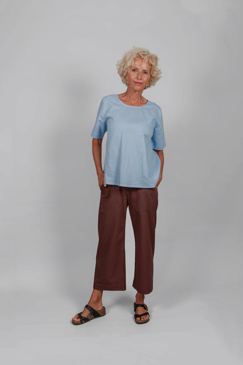 An older woman stands against a light gray studio background wearing a light blue short-sleeve top, dark brown cropped pants, and black sandals.