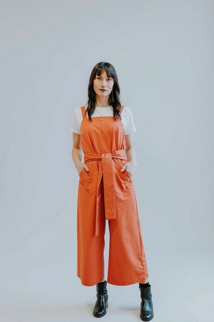 A woman stands against a light gray studio background wearing a bright orange wide-leg jumpsuit over a white short-sleeve top and black ankle boots.