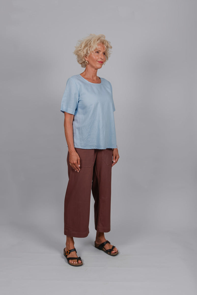 An older woman stands against a light gray studio background wearing a light blue short-sleeve top, brown cropped pants, and black sandals.