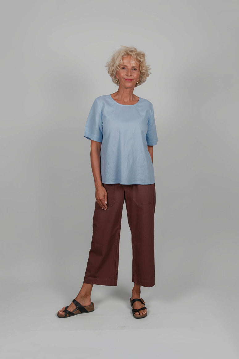 An older woman stands against a light gray studio background wearing a light blue short-sleeve top, dark brown cropped pants, and black sandals.
