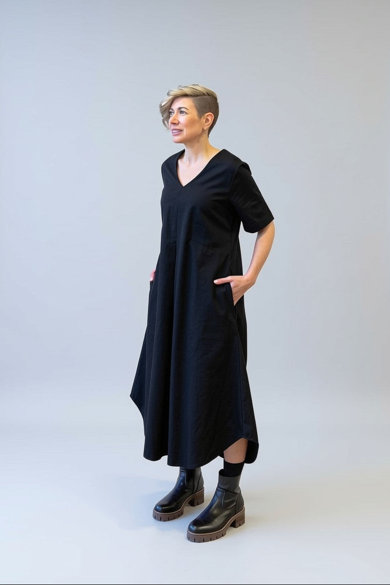 A woman with short blonde hair wearing a loose-fitting black midi dress with short sleeves, black socks, and black leather boots, standing against a light gray studio background.