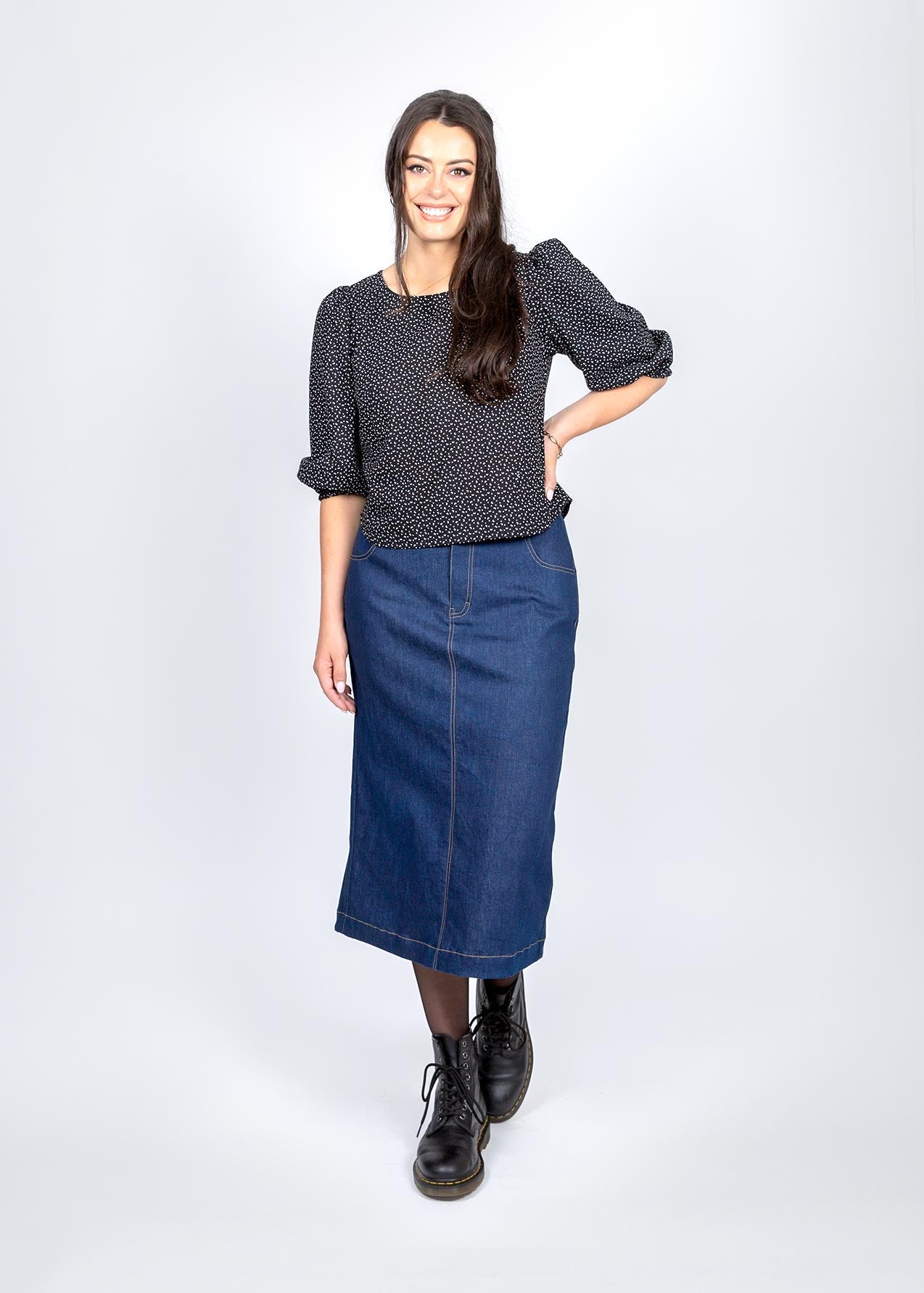 Woman wearing a dark patterned blouse, blue denim skirt, and black boots on a white background
