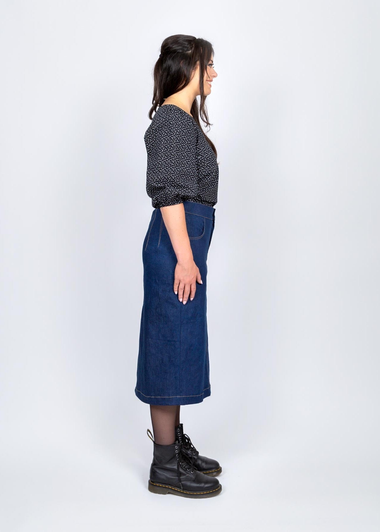 Person wearing a black patterned shirt, blue denim skirt, and black boots on a white background